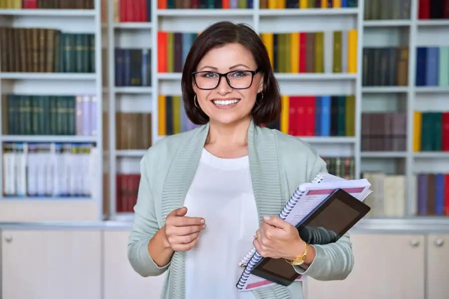 Young smiling woman wearing glasses and professional clothes stands in front several book shelves holding journals and a tablet.