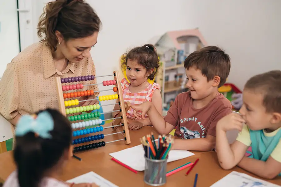 A woman with a ponytail uses a math counter toy as she teaches a group of young children approximately 6 years old in a school setting.