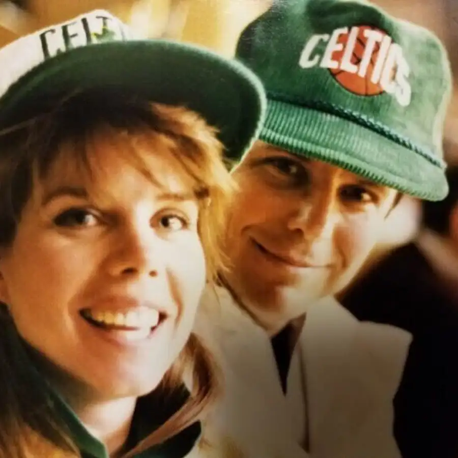 Close-up photo of two smiling people wearing green Boston Celtics hats, with one person in the foreground and the other slightly behind, both appearing happy and cheerful.