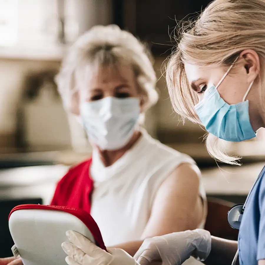 Healthcare worker in a mask and gloves assisting an elderly woman, also wearing a mask, with a medical device in a home setting.