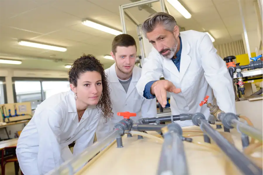 2 young adults in white lab coats are being taught by an older adult male in a white lab coat. They are looking at a plumbing project with pipes and knobs. 