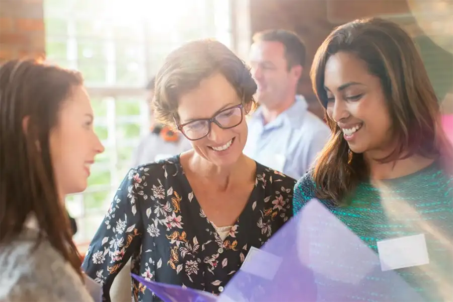 Older woman, wearing glasses, smiling, looks at a folder two younger women are holding for her.