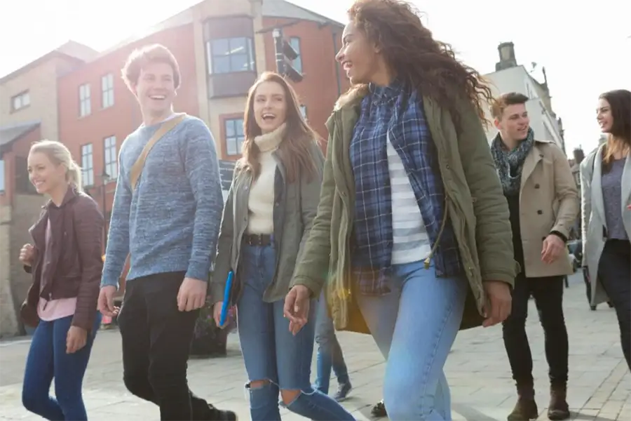 Group of teenagers and young adults are outside engaging with one another, laughing, wearing backpacks, with some holding books.