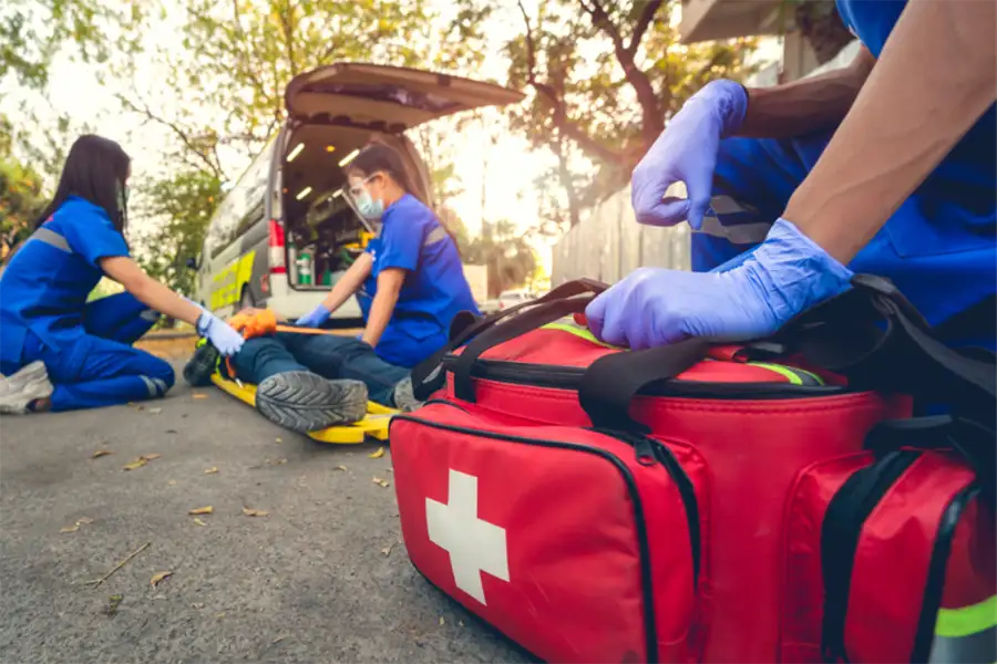 A person is on a stretcher, on the ground, outside of an emergency vehicle as 3 medical workers tend to them. A medical bag is at the forefront of the picture.