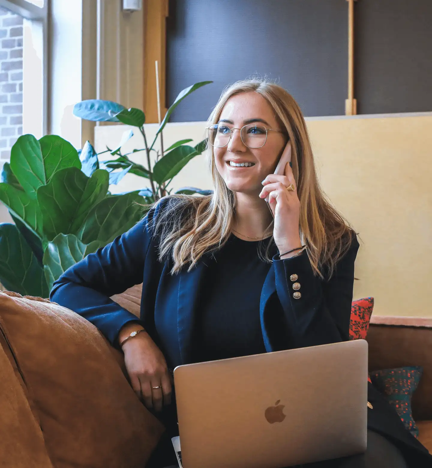 Professional woman with blonde hair, wearing glasses and a dark blazer, sitting on a couch while talking on her phone and smiling, with a laptop on her lap and a large green plant in the background.